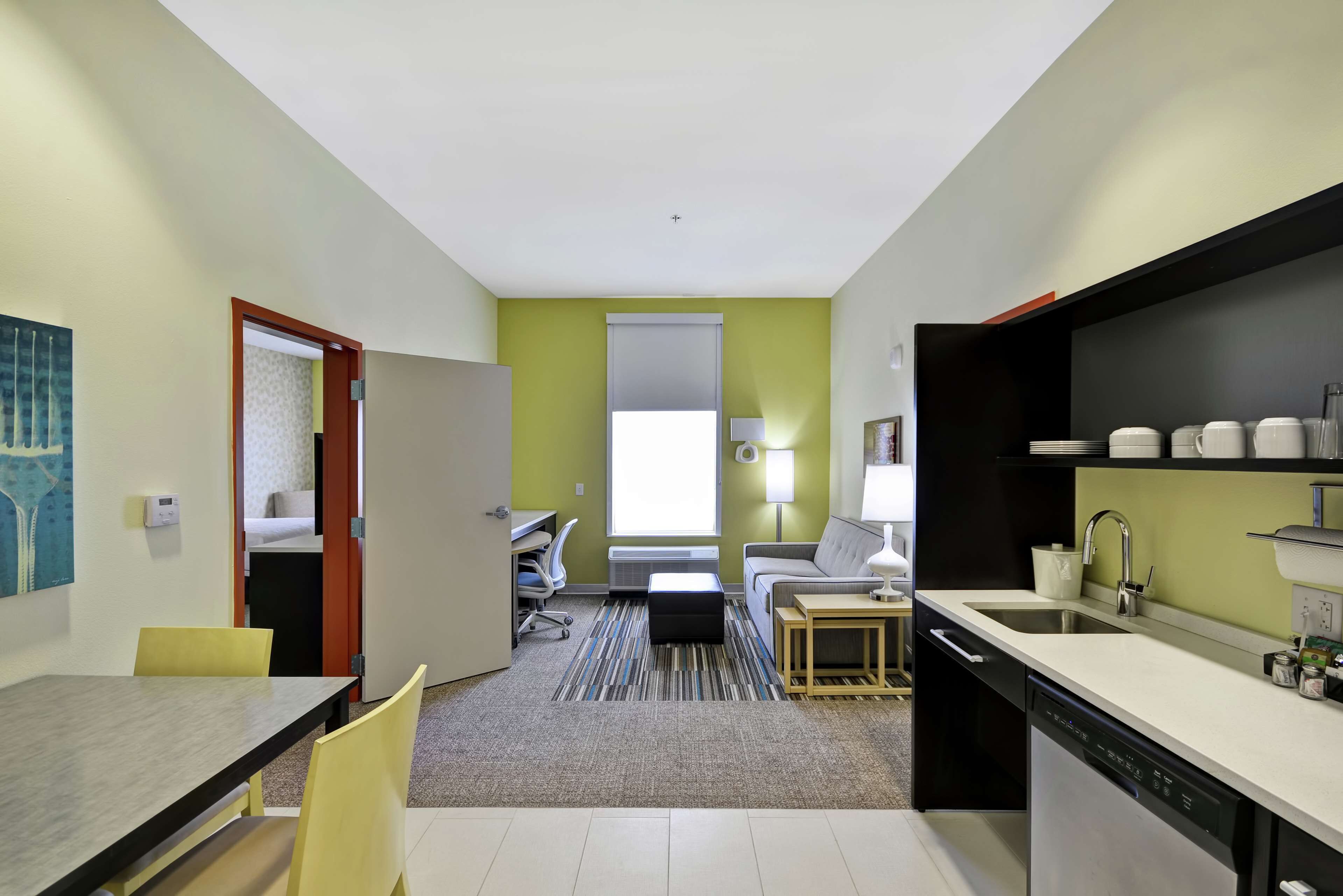 A compact hotel room with a dining area, kitchenette, and a view of the adjacent room.