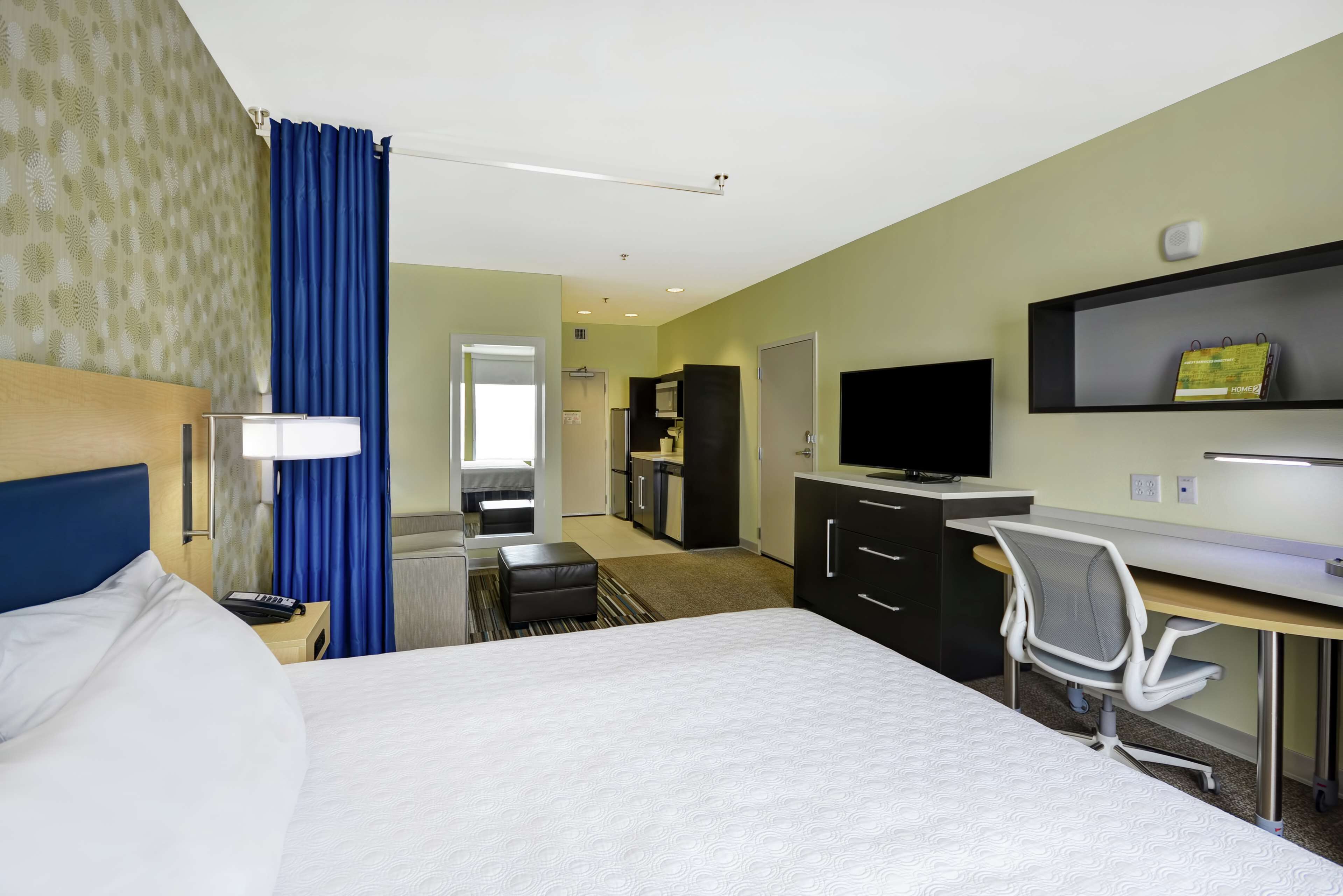 A hotel room with a bed, a desk, a chair, a television, and a kitchenette in the background.