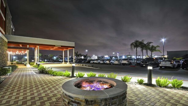 A fire pit area with a stone fire pit, a paved walkway, and a parking lot with cars and palm trees in the background. - Home2 Suites by Hilton Brownsville (Brownsville)