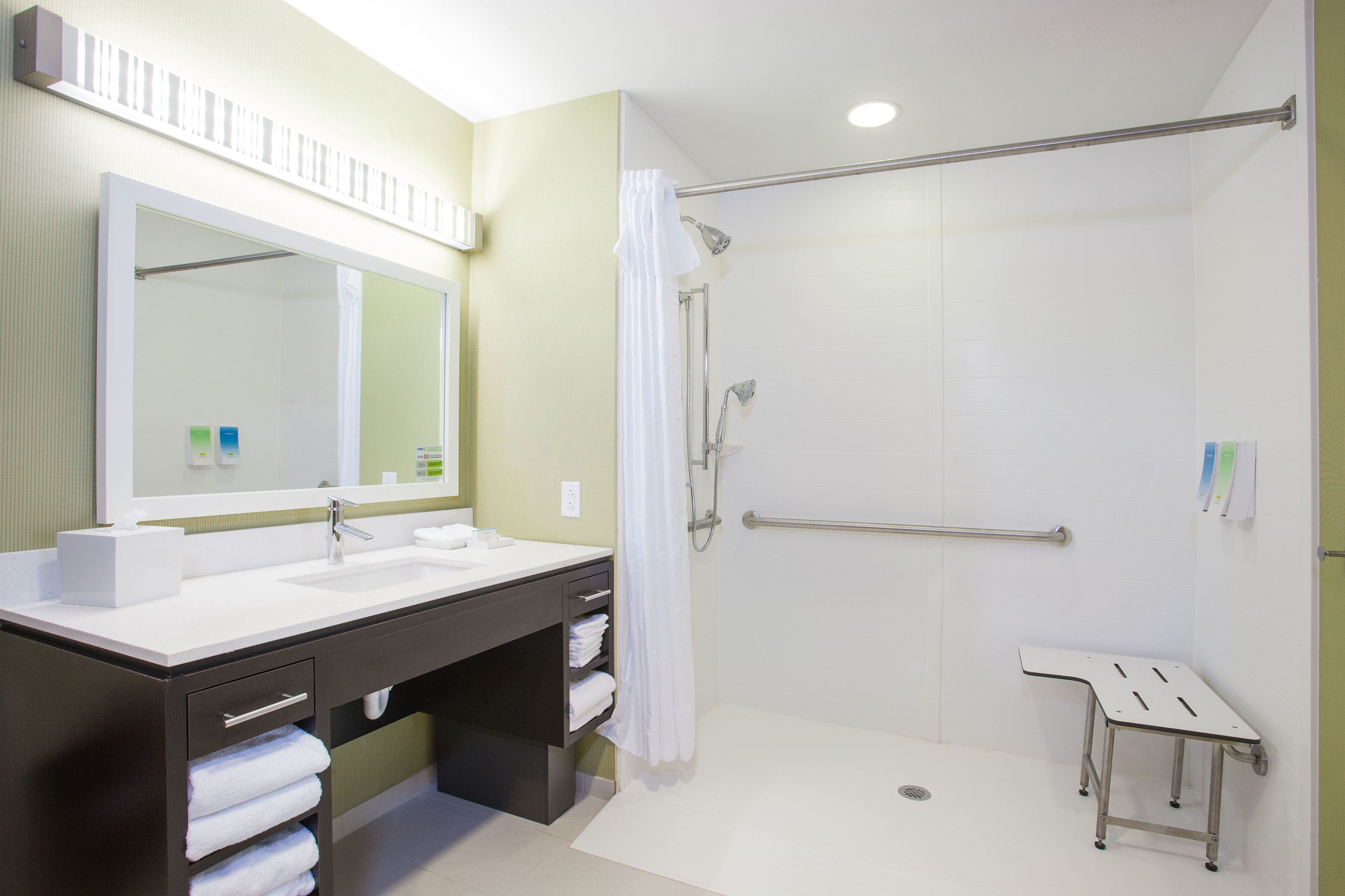 Une salle de bain moderne avec un grand miroir, deux vasques, une douche équipée d’une barre d’appui et une petite table.