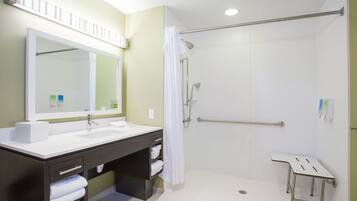 Une salle de bain moderne avec un grand miroir, deux vasques, une douche équipée d’une barre d’appui et une petite table.