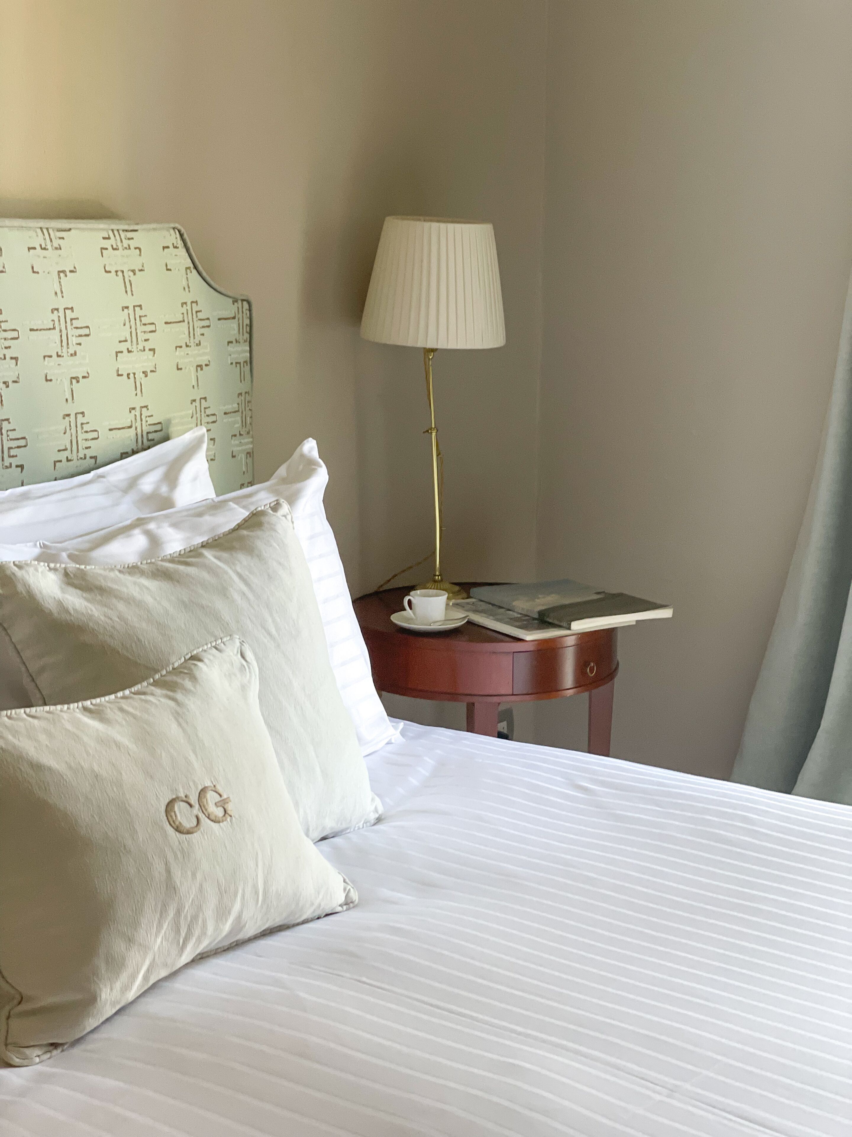 A hotel room with a bed, pillows, a bedside table with a lamp, a cup, and a book.