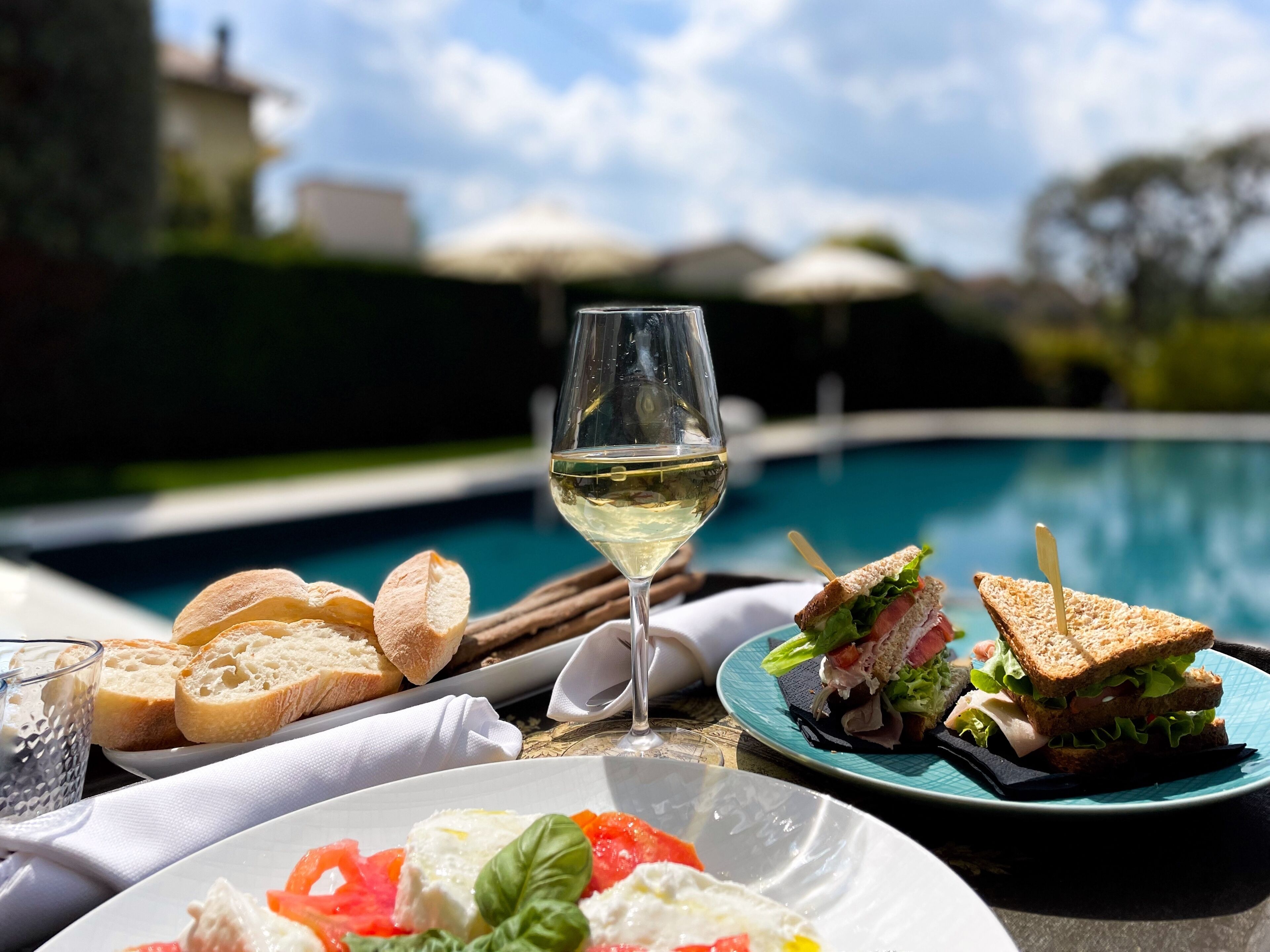 Una mesa de comedor con una copa de vino blanco, sándwiches, pan y un plato de comida junto a una piscina.