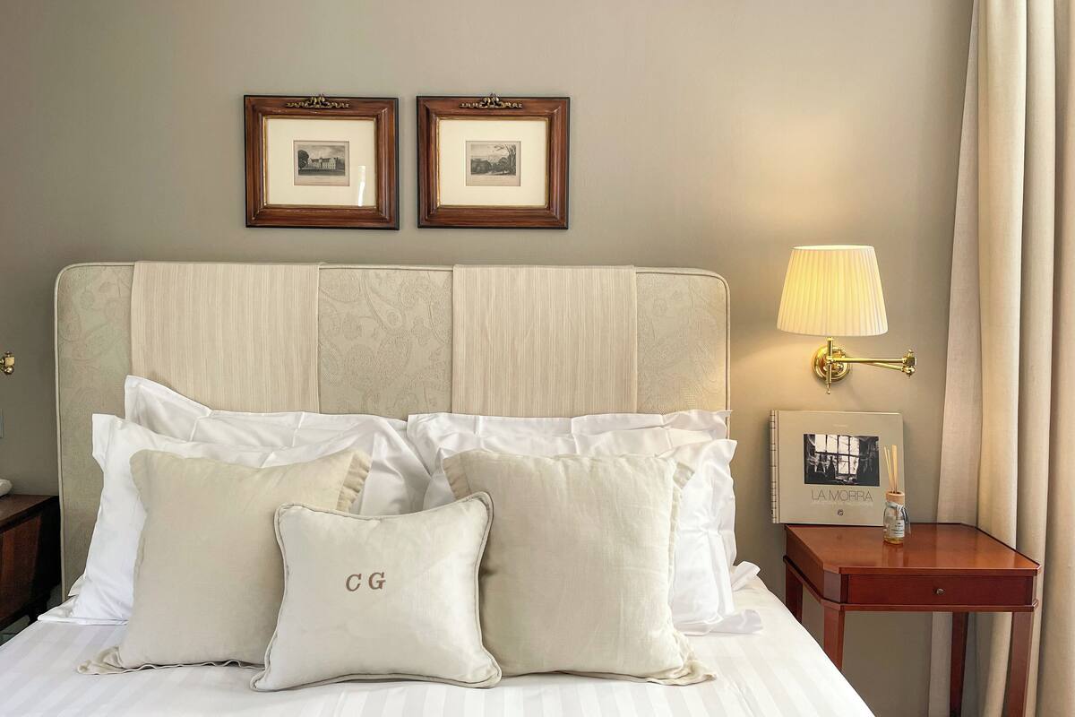 A neatly made bed with white linens and two pillows, one with 'CG' embroidered, a bedside table with a lamp and a picture frame, and two framed pictures on the wall.
