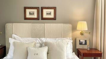 A neatly made bed with white linens and two pillows, one with 'CG' embroidered, a bedside table with a lamp and a picture frame, and two framed pictures on the wall.