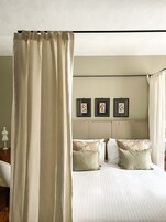 Un dormitorio con cama a dosel, cabecera con tres cuadros enmarcados y mesita de noche con lámpara.