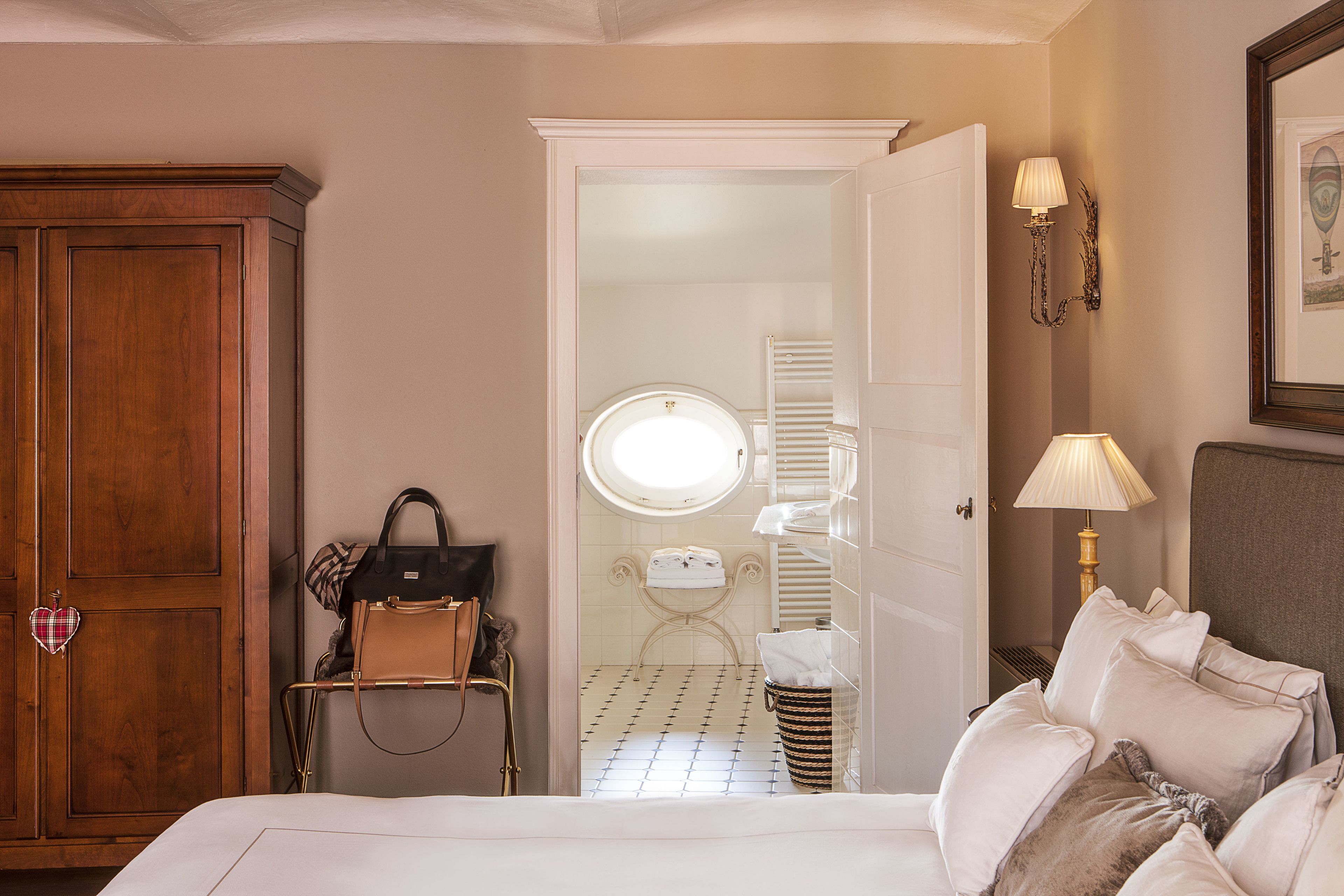 Une chambre d’hôtel avec une grande armoire en bois, un lit avec plusieurs oreillers, un miroir rond, une petite salle de bain avec des toilettes et un porte-serviettes, et une lampe fixée au mur.