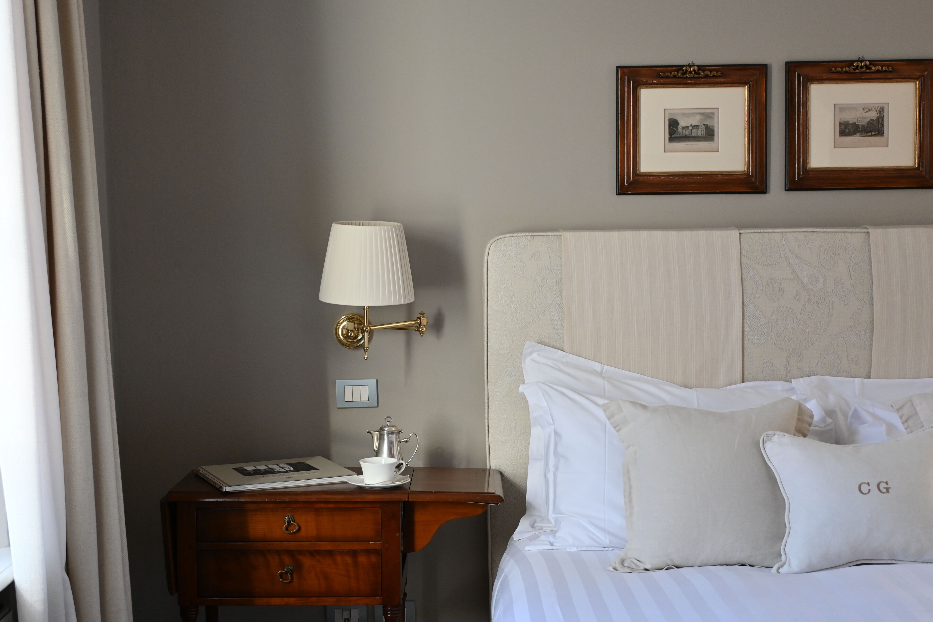Una habitación de hotel con una cama, una mesita de noche con una lámpara, un cuadro enmarco y una pequeña mesa con una taza y un libro.