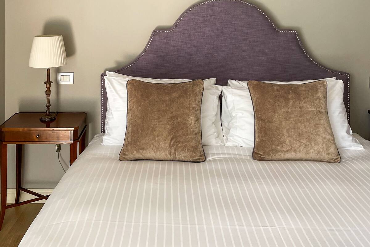 A neatly made bed with striped bedding, a headboard with a purple fabric and decorative bead trim, and a bedside table with a lamp and a power outlet.