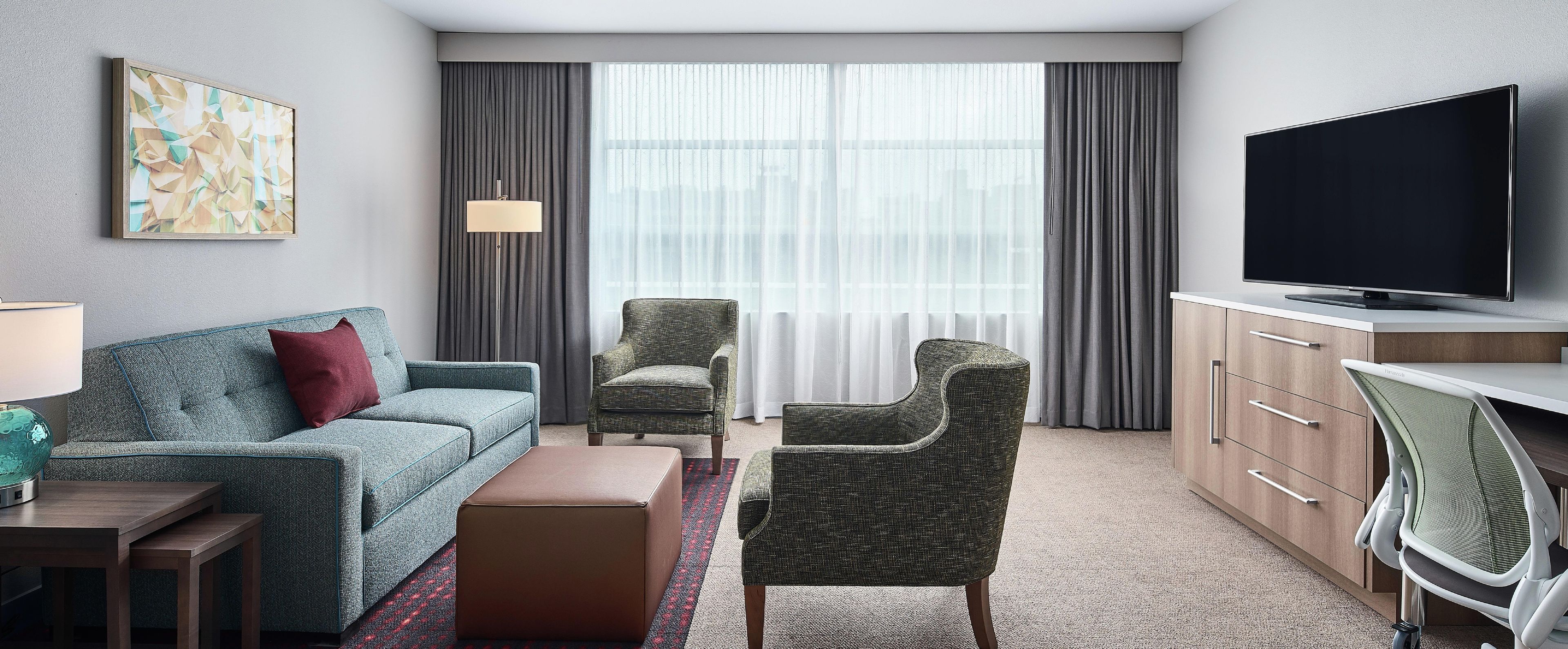 Habitación de hotel con sofá, dos sillones, televisión, escritorio y un ventanal con cortinas.