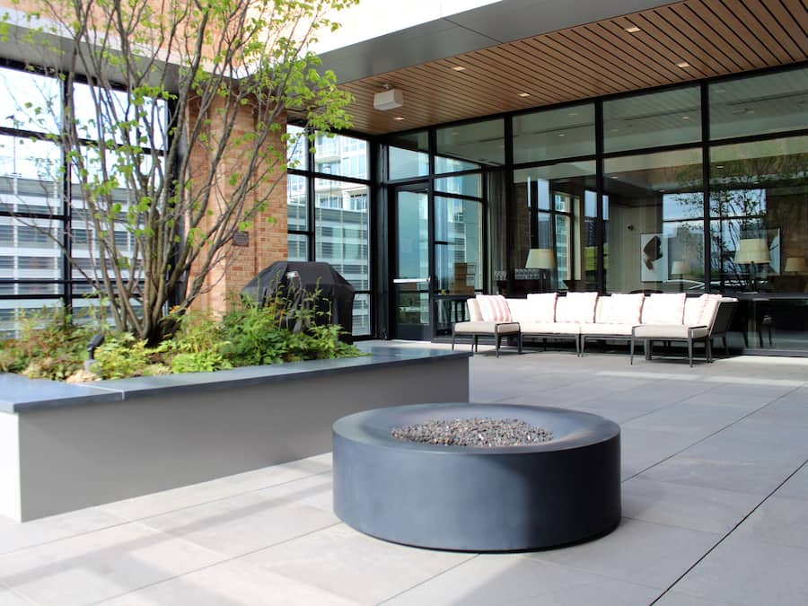 An outdoor seating area with a fire pit, a brick planter with greenery, and a modern building with large windows.