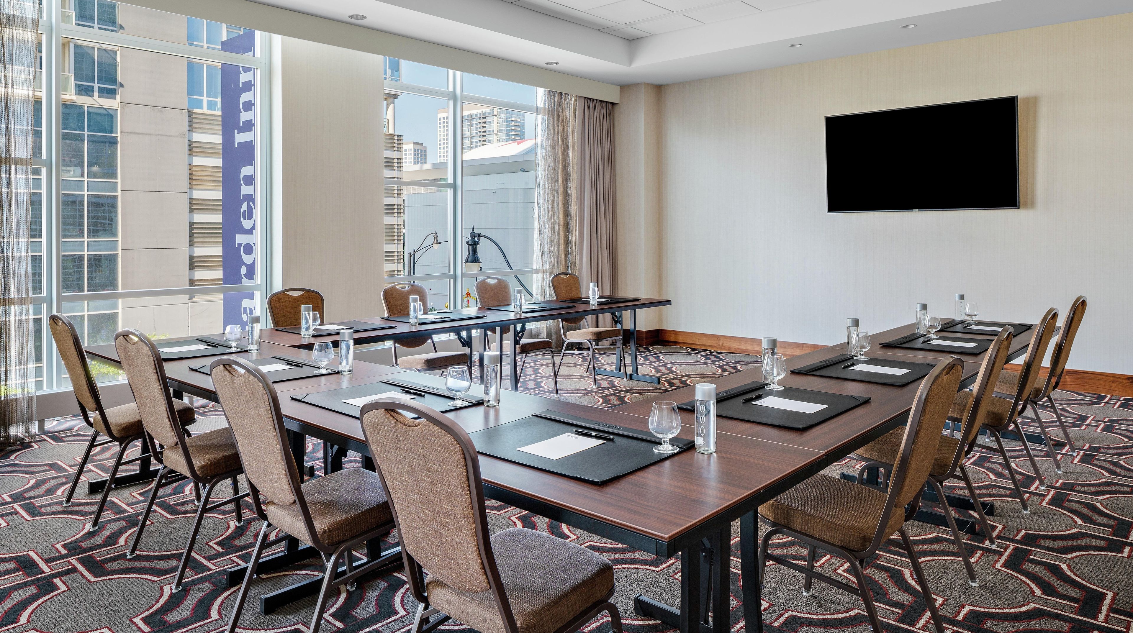 A conference room with a long table, chairs, a flat-screen TV, and large windows with a city view.