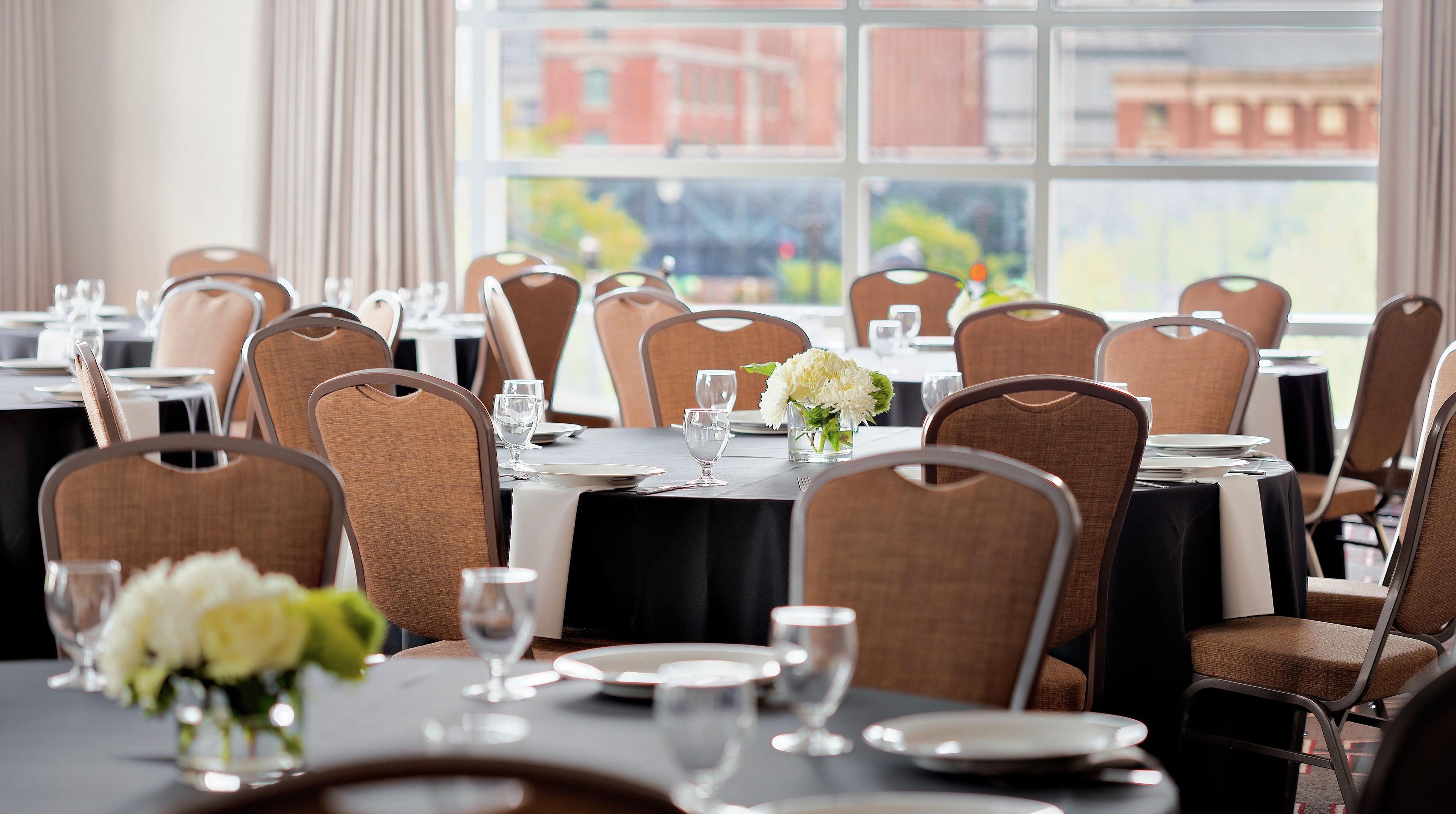 A dining area with tables set for service, chairs, and a view of buildings through large windows.