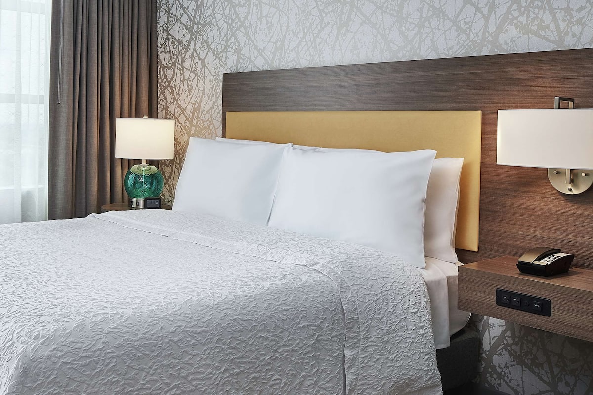 A hotel room with a large bed, two bedside lamps, a nightstand with a phone, and a window with sheer curtains.