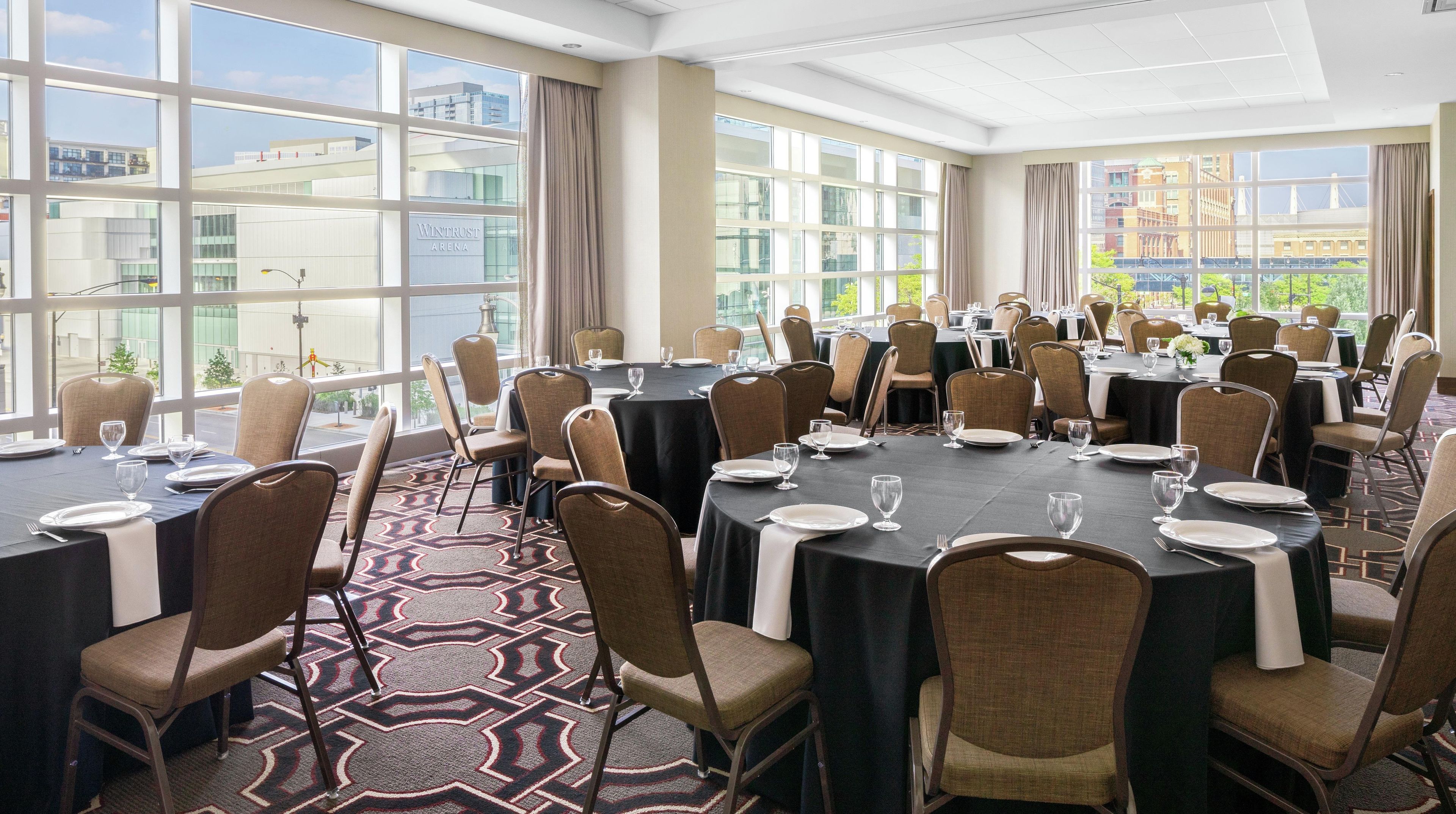 A conference room with multiple tables and chairs, large windows with a city view, and a patterned carpet.