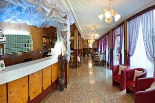 Una hall d'albergo con una reception, un lampadario e sedie rosse.