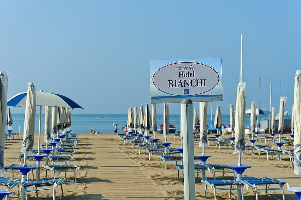 Una spiaggia con file di lettini e ombrelloni blu e bianchi, un cartello con la scritta Hotel Blanchi e l'oceano sullo sfondo.