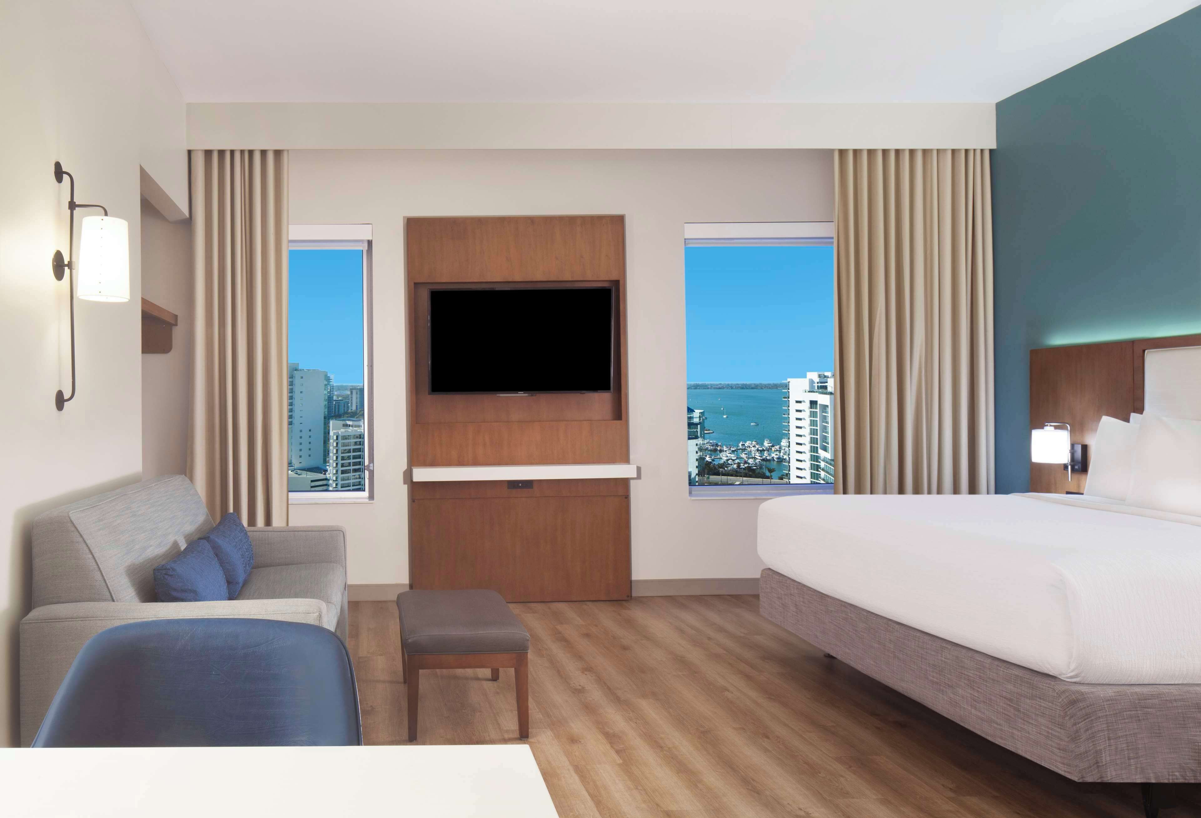 Quarto de hotel com uma cama grande, um sofá, uma TV na parede e uma janela com vista para a cidade.