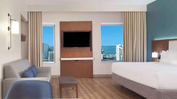 Quarto de hotel com uma cama grande, um sofá, uma TV na parede e uma janela com vista para a cidade.