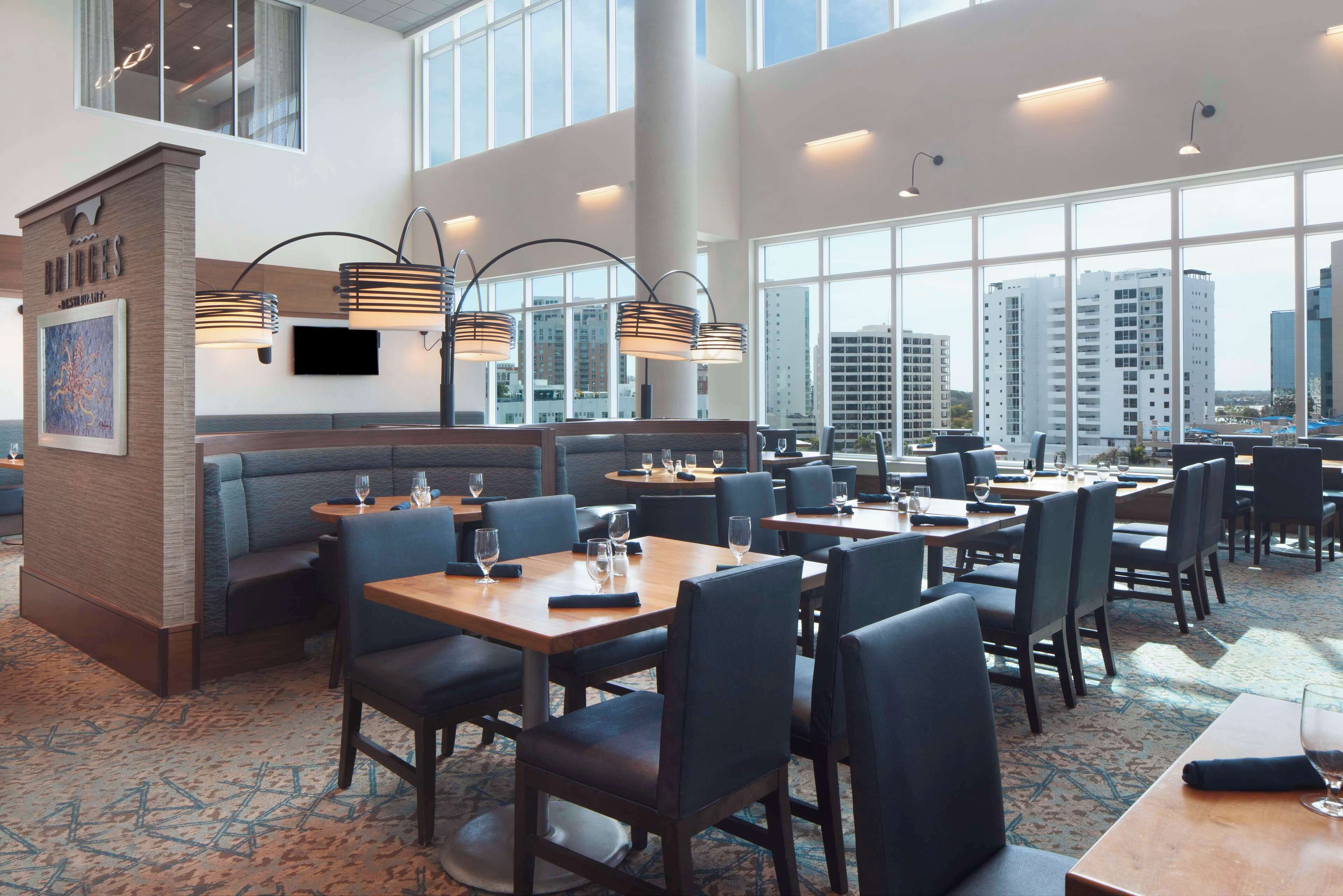 Un comedor de hotel moderno con techos altos, ventanas amplias y una variedad de opciones de asiento, incluyendo mesas y cabinas.