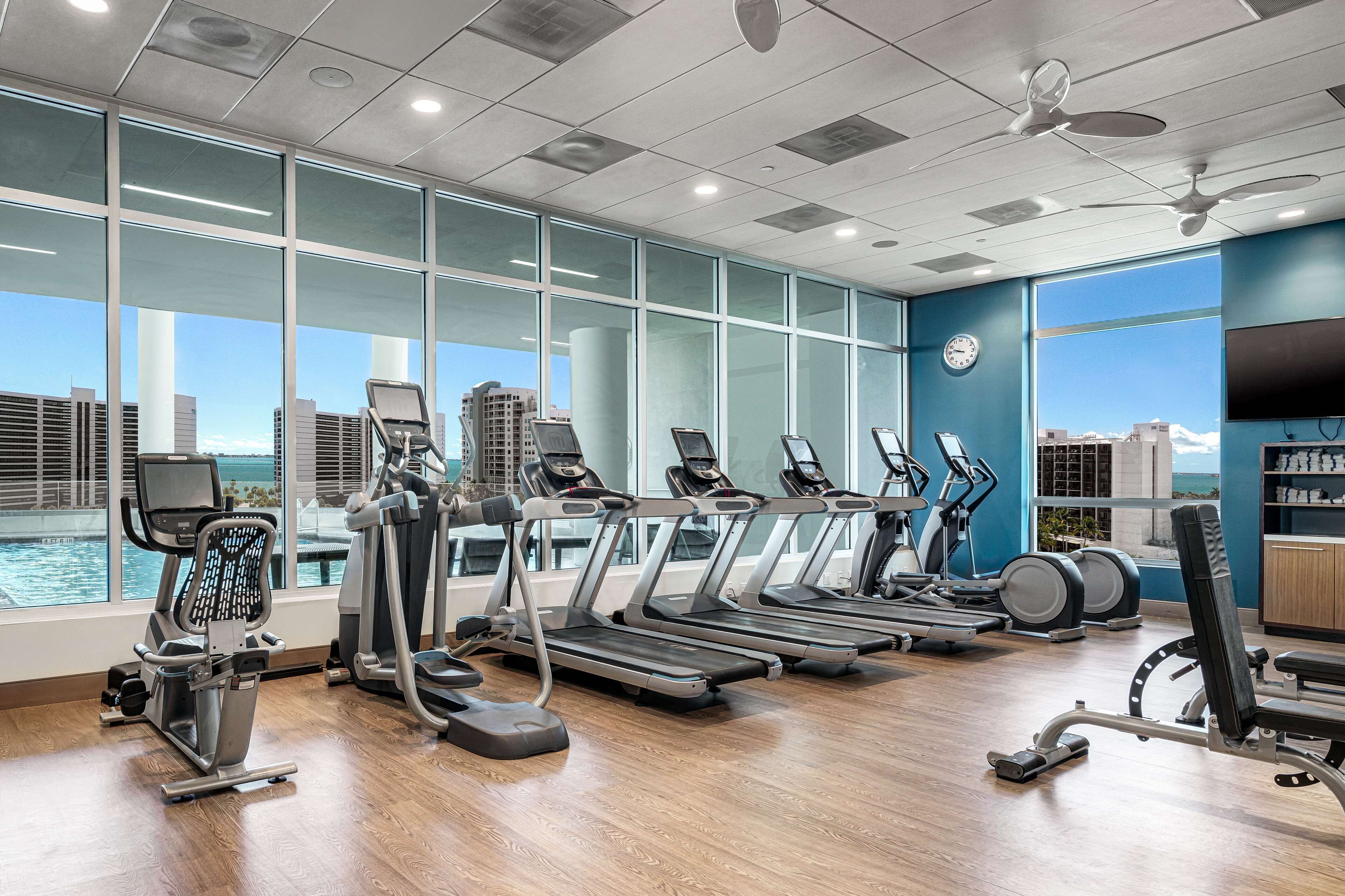 A gym with treadmills, ellipticals, and a rowing machine, large windows with a city view, and a ceiling fan.