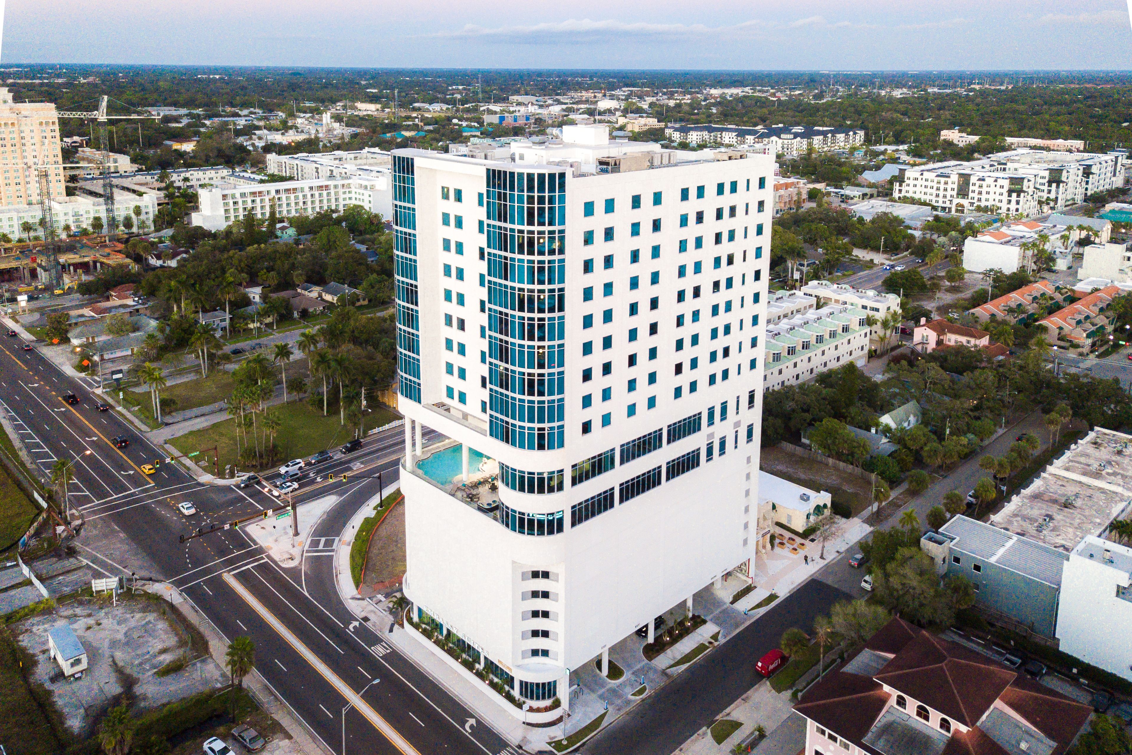 A multi-story hotel with a modern design, featuring a prominent corner location, a rooftop pool, and a view of the surrounding cityscape.