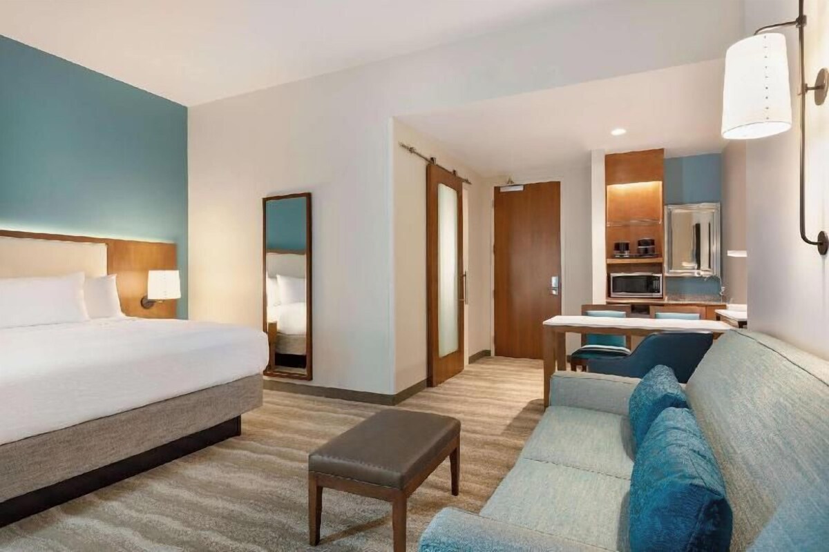 A hotel room with a bed, a sofa, a small table, a mirror, and a kitchenette area.
