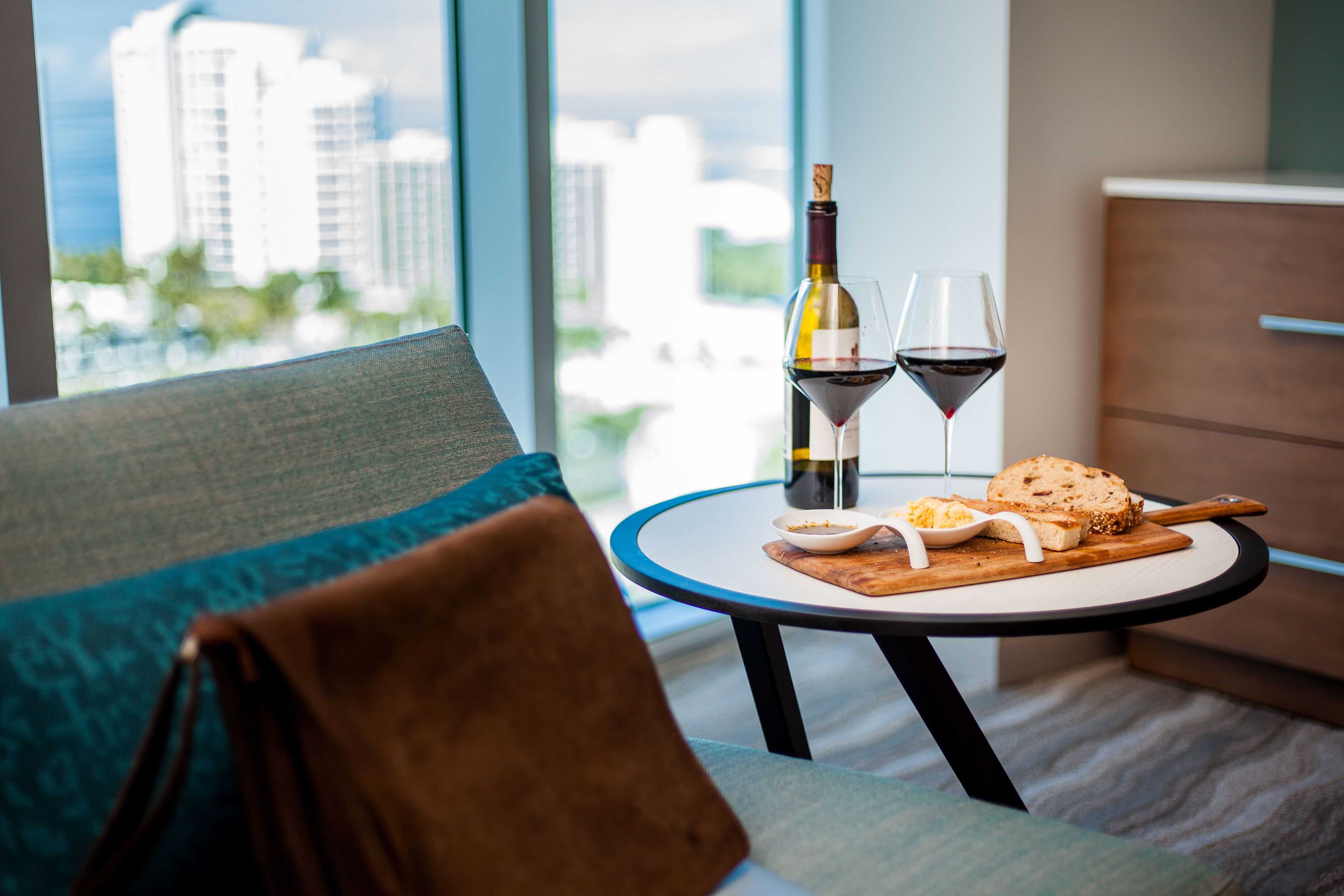 A modern living room with a view of a cityscape through large windows. A round table with a bottle of wine, two glasses, and a plate of food is placed in the center.