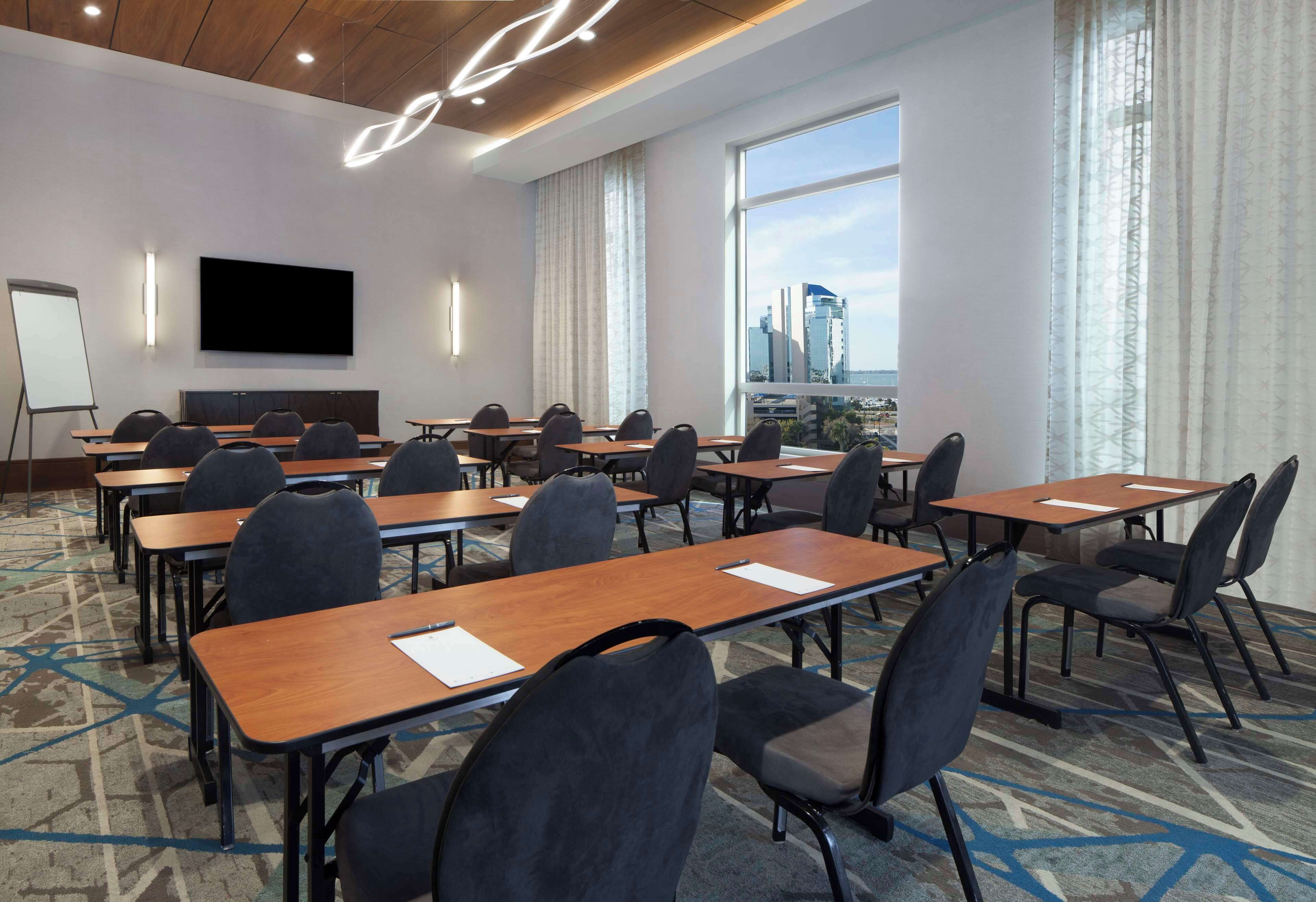 A conference room with multiple tables and chairs, a large screen, and a view of a cityscape through large windows.