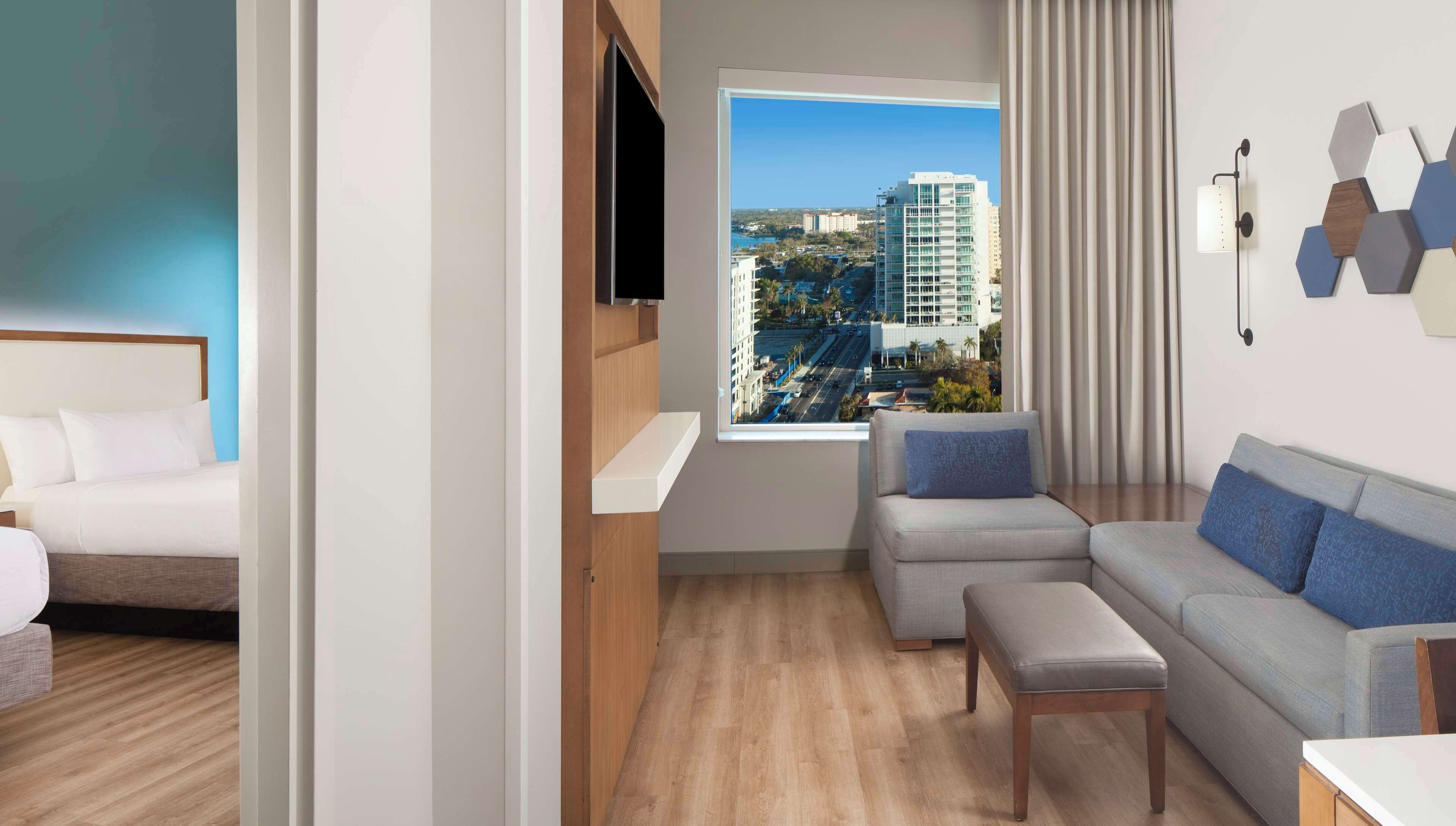 A modern hotel room with a bed, a sofa, a bench, and a large window offering a city view.