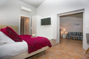 A bedroom with a bed, a bedside table, an air conditioner, a wardrobe, a television, a sign, and a living area with a sofa and a lamp. - Le Relais Des Trois Mas (COLLIOURE)
