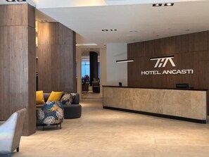 Zona de recepción de un hotel, con mostrador de recepción, un sofá con cojines y una pared de madera con el nombre del hotel.