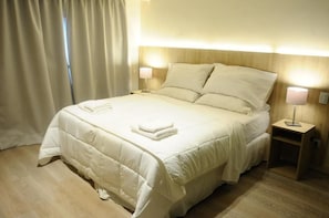 Una habitación de hotel con cama, dos lámparas de noche, mesita de noche y ventana con cortinas.