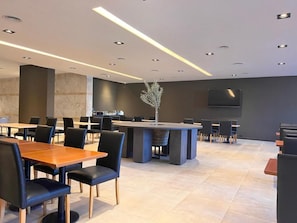 A modern dining area with multiple tables and chairs, a central decorative plant, and a flat-screen TV mounted on the wall.