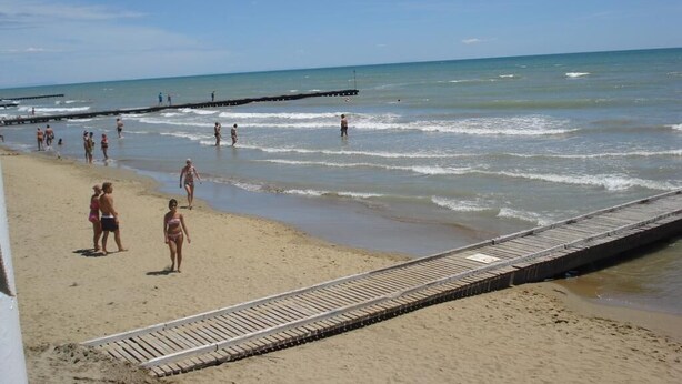 A beach with a wooden boardwalk, sandy shore, and people walking along the water. - Hotel Roby (Jesolo)