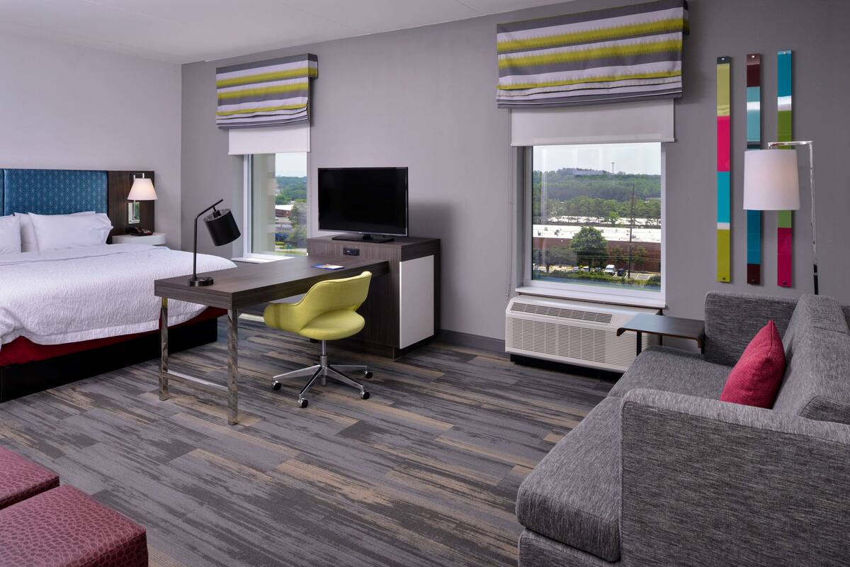 A hotel room with a bed, a desk with a TV, a yellow chair, a grey sofa, and a view of the city.