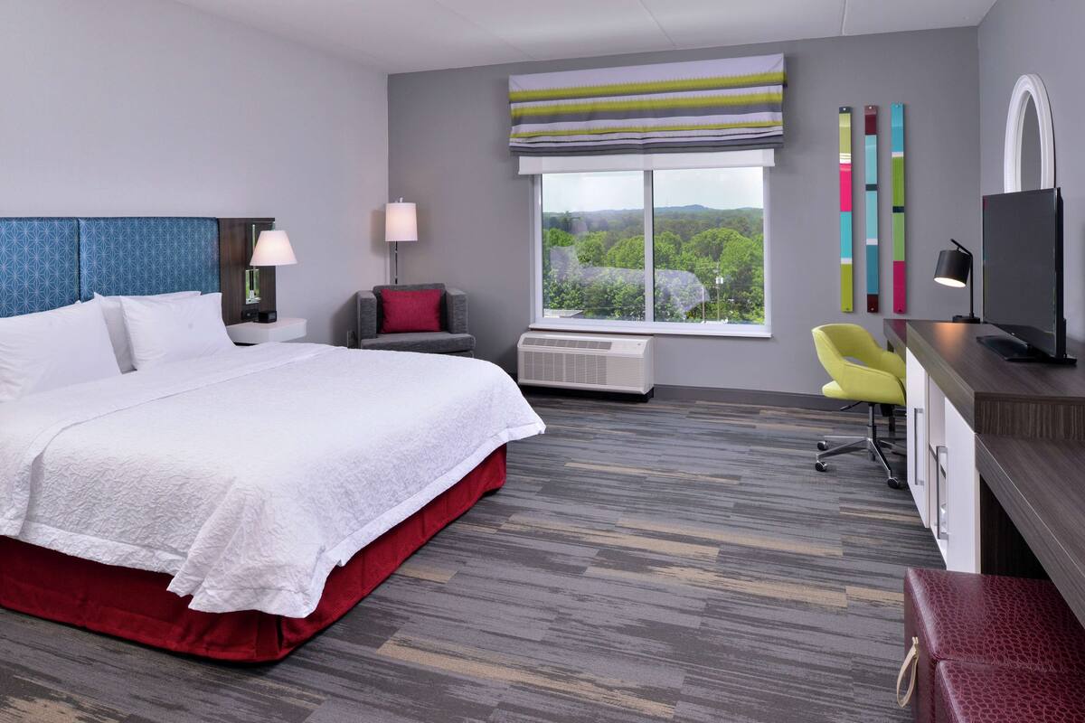A modern hotel room with a large bed, a desk with a computer, a chair, and a view of greenery outside.