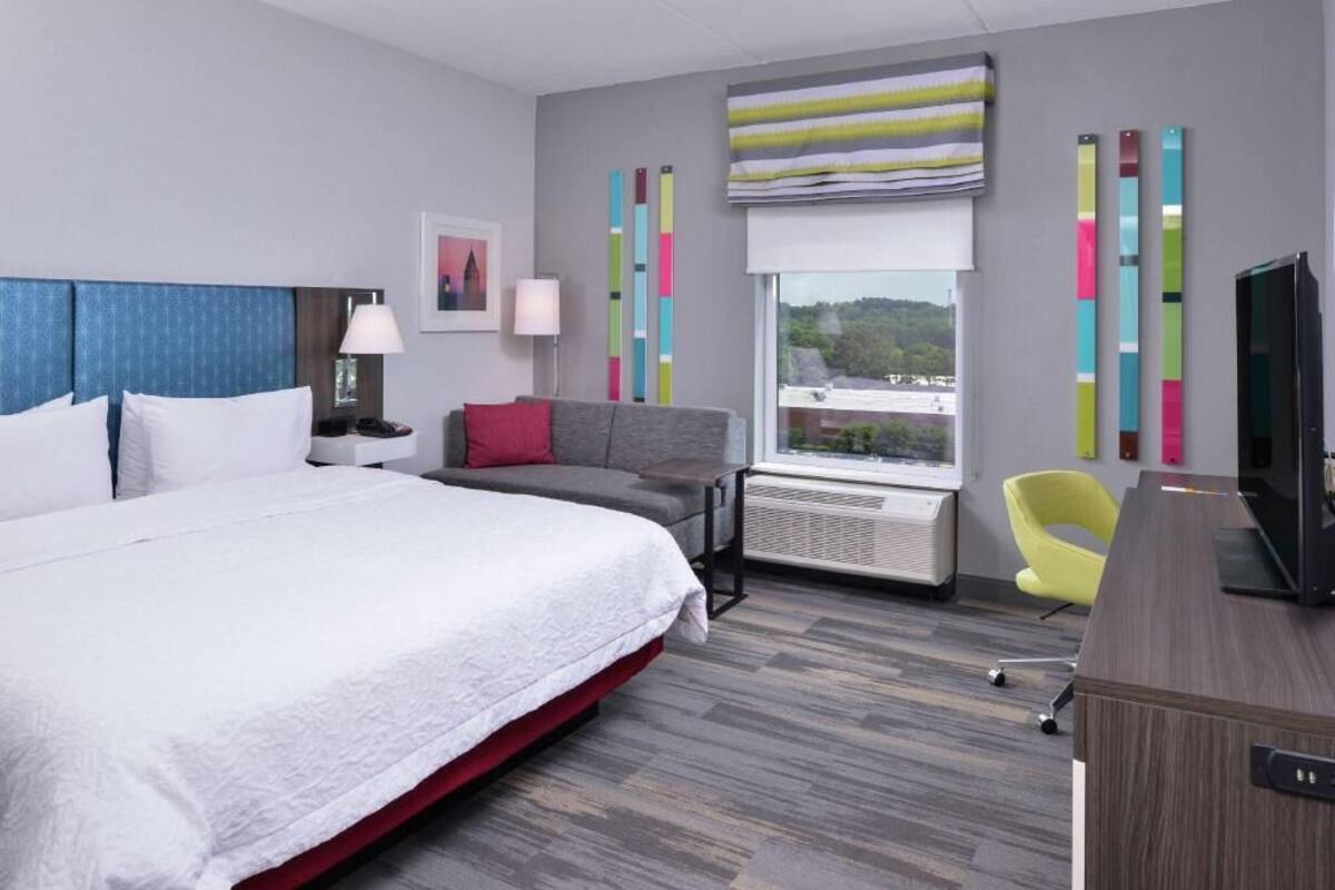 A modern hotel room with a large bed, a desk, a chair, a television, and a window with a view.