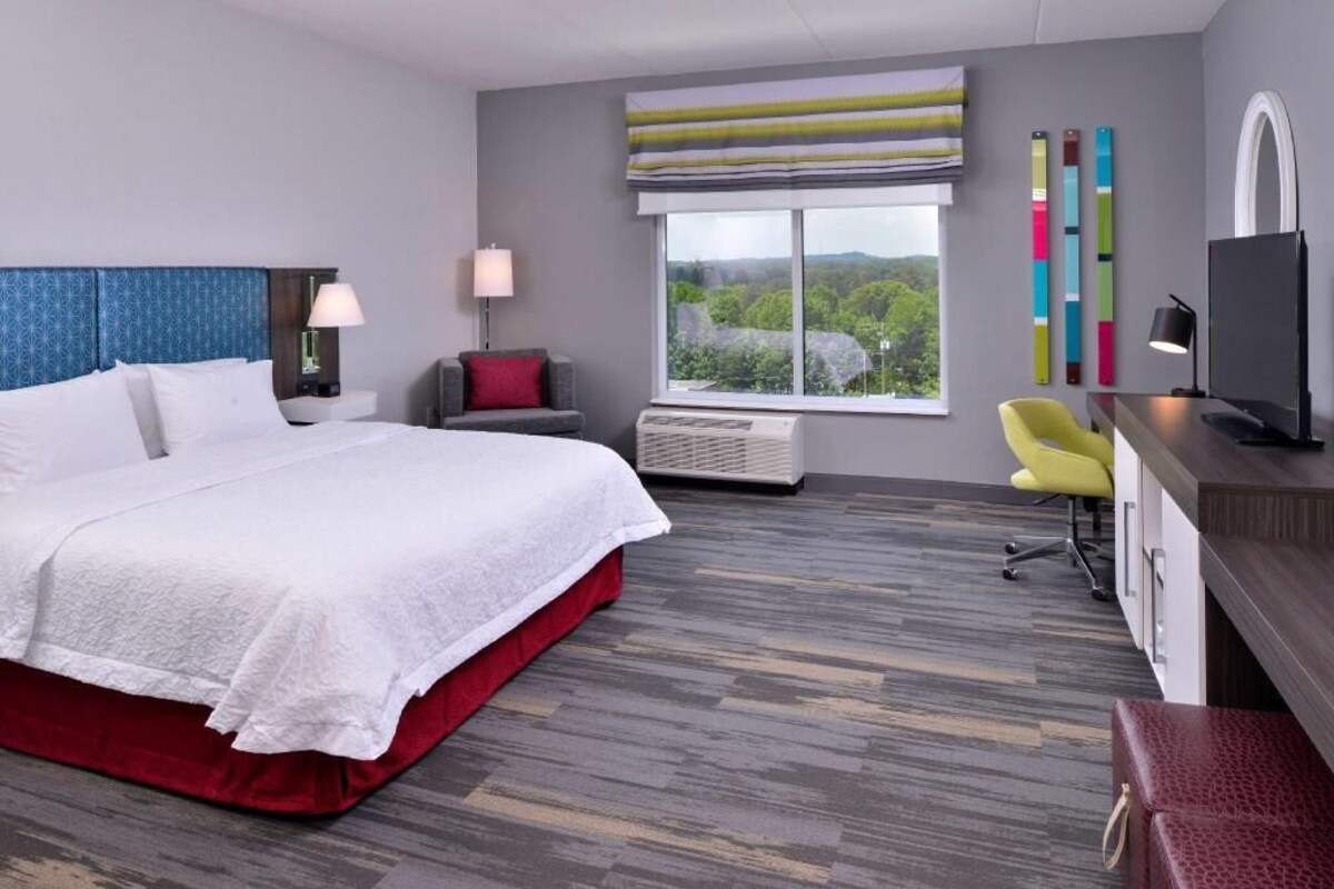 A modern hotel room with a large bed, a desk with a computer, a chair, a window with a view, and a TV.