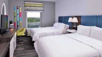 A hotel room with two beds, a desk with a chair, a TV, and a window with a view of greenery.