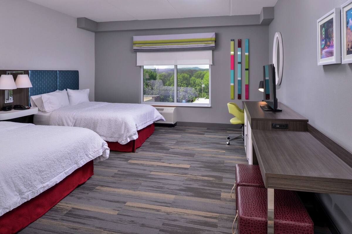 A hotel room with two beds, a desk, a chair, and a window with a view of greenery.