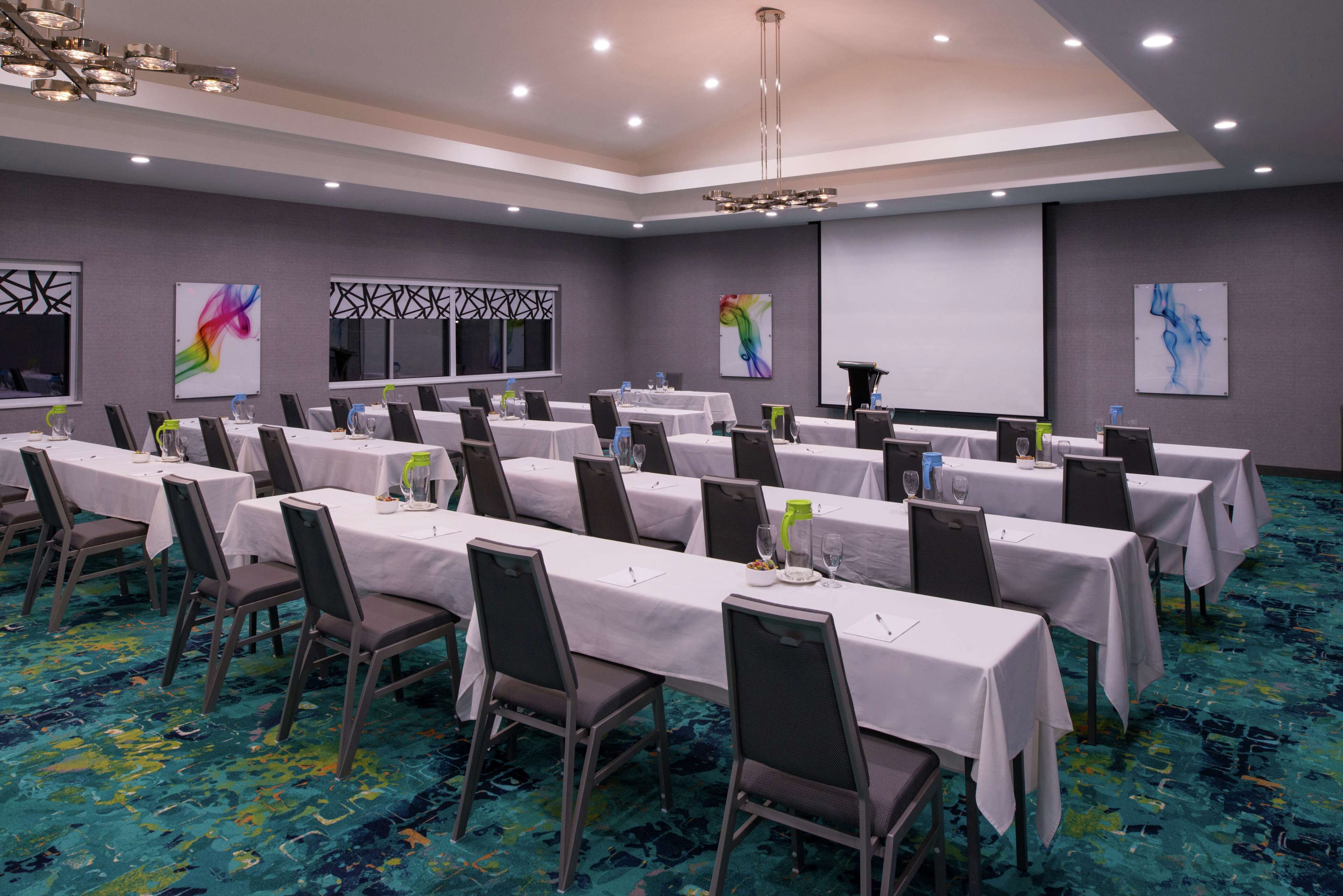 A conference room with multiple tables and chairs, equipped with water bottles and glasses, and a projector screen on the wall.
