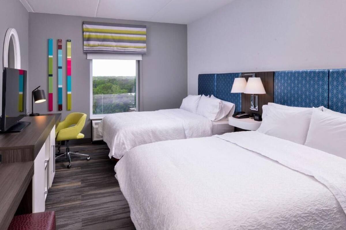 A hotel room with two beds, a desk with a chair, a window with a view, and a lamp.