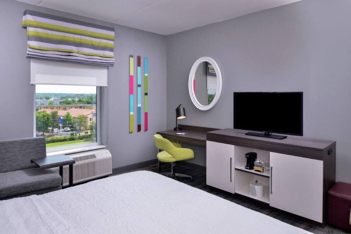 A modern hotel room with a bed, a desk with a chair, a television, a window with blinds, and a view of the outside.