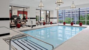 A modern indoor swimming pool with clear glass walls, a seating area with chairs and red vases, and large windows offering a view of the city. - Hilton Garden Inn Chicago McCormick Place (Chicago)