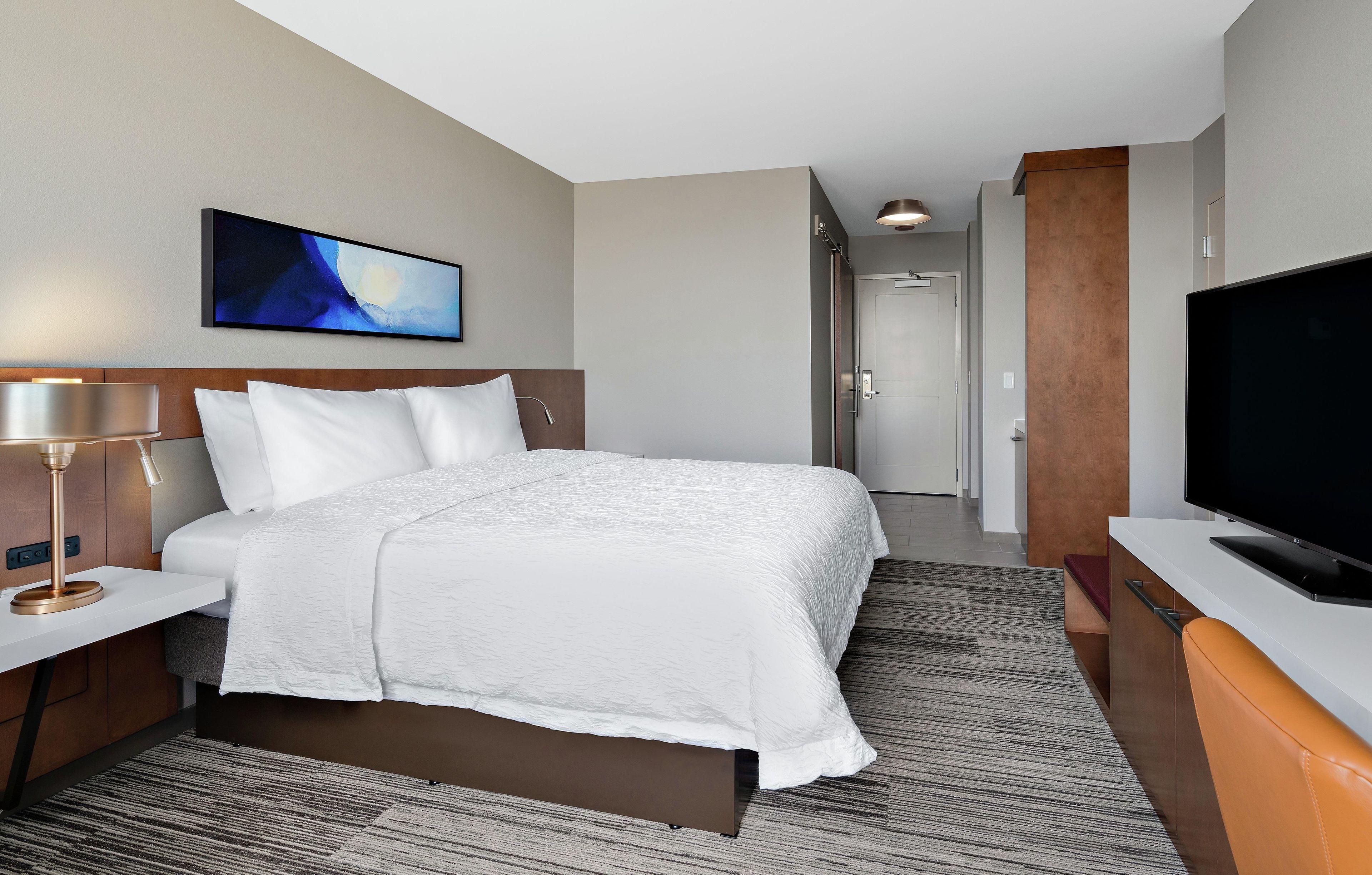 A hotel room with a bed, a TV, a desk, and a painting on the wall.