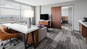 A hotel room with a large window, a desk with a lamp, a sofa, a TV, and a kitchenette.