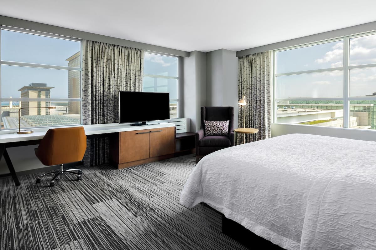 Une chambre d’hôtel avec un grand lit, un bureau avec une télévision, une chaise et une vue sur le paysage urbain grâce à de grandes fenêtres.