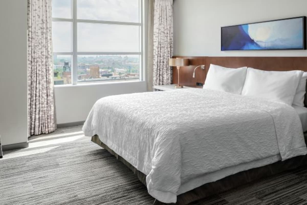 A hotel room with a large bed, a nightstand, a lamp, and a view of the city through a window.