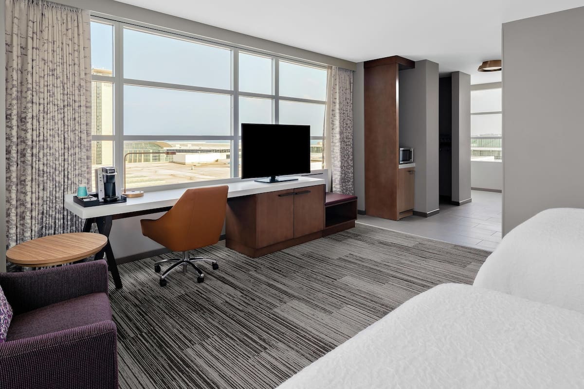 Une chambre d’hôtel avec une grande fenêtre, un bureau avec un ordinateur, une chaise, un lit et une petite table.
